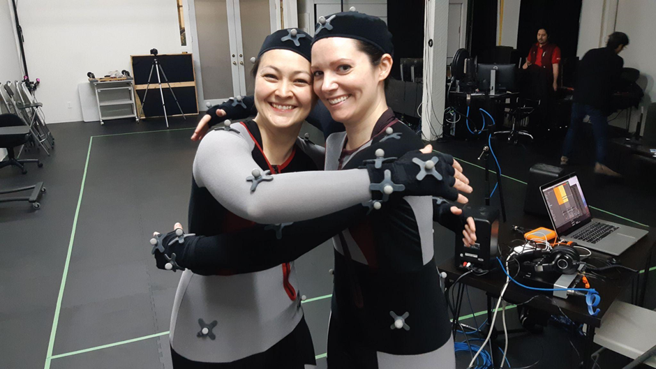 Photo by Brian Topp. Photo of Debi Wong and Mireille Asselin embracing one another and smiling facing the camera while wearing motion capture suits in a studio with computer equipment and artists in the background. There are boxes marked out in green tape on the floor.